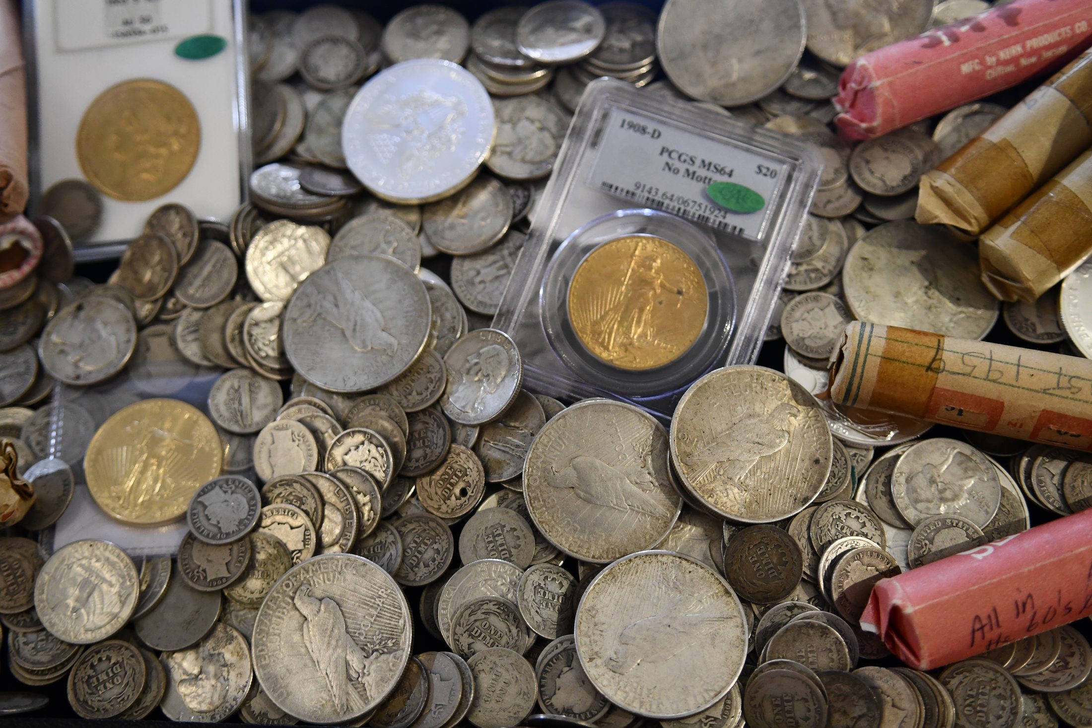 Dealer table covered in raw silver dollars and dimes, original coin rolls, and a certified 1908-D twenty-dollar gold piece